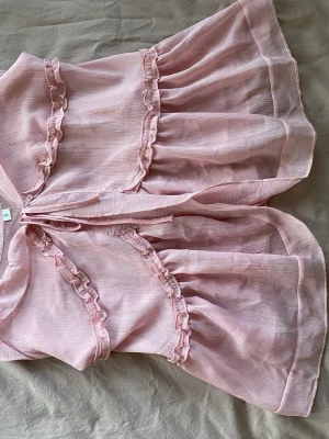 Ljusrosa bolerotopp med volanger - Supersöt ljusrosa bolerotopp i transparent polyester med volangdetaljer och knyt framtill. Perfekt för att lägga till en romantisk touch till din outfit. Luftig och flowy modell med korta ärmar.