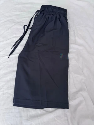 Mörkgrå shorts från Under Armour - Snygga mörkgrå shorts från Under Armour med elastisk midja och snörning. Tillverkade i ett lätt och bekvämt syntetmaterial, perfekta för träning eller chill. Diskret logga på benet och enkel, clean design.