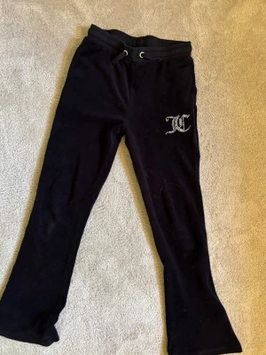 Svarta mjukisbyxor Juicy Couture strl 10/11 år - Mörka, mjuka velourbyxor från Juicy Couture med dragsko i midjan och två bakfickor. Byxorna har ett glittrigt JC-logotyptryck på ena benet och utsvängda ben. Perfekta för chill dagar eller när du vill vara extra comfy. De är ett hål vid fickan längst ner men de syns inte men man kan inte ha grejer i den fickan för då ramlar de ut.