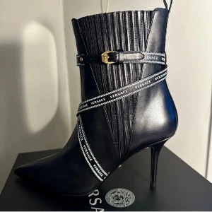 Svarta stövletter från Versace - Snygga svarta stövletter från Versace i läder. Dekorativa band med Versace-logga och guldfärgat spänne ger en exklusiv look. Stövletterna har ribbade detaljer på skaftet och dragögla bak. Oanvänd stl 39 liten 40. Nypris 11.299:- (NK huset) 