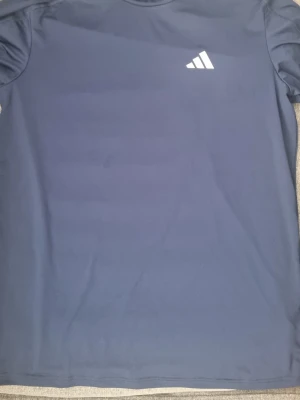 Blå tränings-tshirt från Adidas - Snygg blå tränings-tshirt från Adidas med klassisk vit logga på bröstet. Tillverkad i ett lätt och snabbtorkande material som passar perfekt för gymmet eller löprundan. Enkel design med rund hals och korta ärmar.