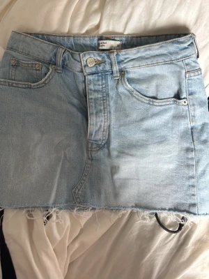 Ljusblå jeanskjol med fransig kant - Säljer en ljusblå jeanskjol från Perfect Jeans med klassisk femficksdesign och rå, fransig kant nedtill. Kjolen har bälteshällor och knappgylf, perfekt för en avslappnad och trendig look.