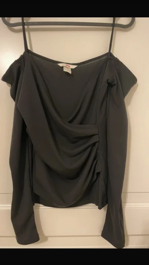 Svart off-shoulder tröja - Trendig svart omlottopp med draperad framsida och tunna axelband. Toppen har långa ärmar och en snygg offshoulder-look som ger en cool vibe. Perfekt för dig som gillar stilrena plagg med en twist.