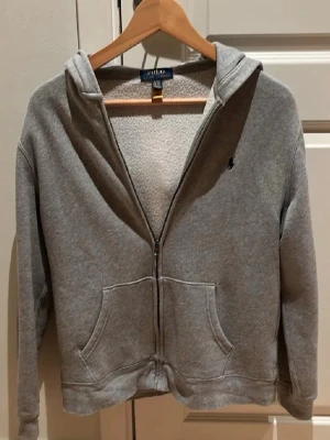 Grå hoodie från Ralph Lauren XS - Grå hoodie från Polo Ralph Lauren i storlek XL barn / xs/ S i vuxen med dragkedja och huva. Tröjan har två stora fickor framtill och ett litet broderat logomärke på bröstet. Insidan är mjuk och fleecefodrad, perfekt för kyliga dagar. Klassisk och enkel design som funkar till allt.