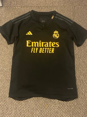 Svart Real Madrid tränings t-shirt - Snygg svart tränings t-shirt från Adidas med Real Madrid-logga och gula detaljer. Framsidan har trycket 'Emirates Fly Better' i gult och tre diskreta ränder på axlarna. Perfekt för fotbollsträning eller gymmet.