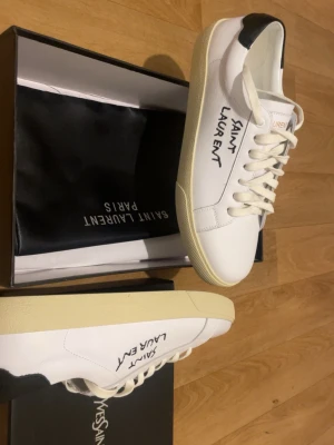 Saint Laurent vita sneakers - Snygga vita sneakers från Saint Laurent med svart detalj på hälen och handskriven logga på sidan. Skorna har klassisk låg profil, vita snören och sula i offwhite. Tillverkade i läder för en clean och exklusiv look.Helt oanvända.
