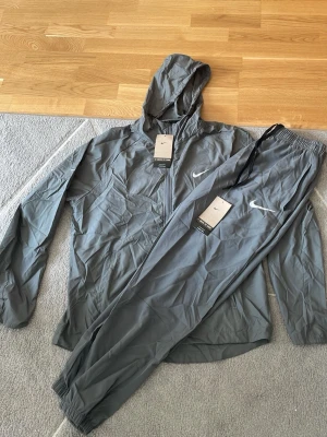 Nike Miller grå  - Snyggt grått träningsset från Nike med jacka och byxor. Jackan har huva, dragkedja och två fickor framtill. Byxorna har resår i midjan och vid benslut samt dragsko. Båda plaggen har Nike-logga på bröst och lår. Perfekt för träning eller chill.