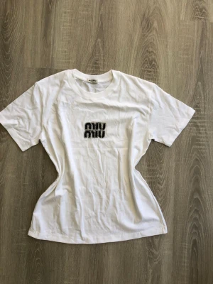 Vit Miu Miu t-shirt med tryck - Säljer en vit t-shirt från Miu Miu med svart tryck på bröstet. T-shirten har klassisk rund halsringning och korta ärmar. Tillverkad i mjuk bomull för en bekväm känsla och enkel stil.
