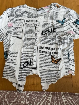 Vit t-shirt med tidningsmönster och fjärilar - Unik vit t-shirt med tidningsinspirerat tryck, blå och orange fjärilar samt rosa rosor. T-shirten har korta ärmar och en croppad, ojämn nederkant för en edgy look. Perfekt för dig som vill sticka ut med coola detaljer och grafiskt mönster.