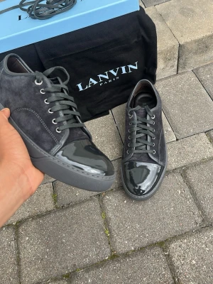 Lanvin svarta sneakers i mocka och lack - Snygga svarta sneakers från Lanvin med ovandel i mocka och glansig lackdetalj på tån. Skorna har gråa skosnören, metallfärgade öljetter och en robust gummisula. Perfekt för dig som gillar exklusiv och stilren design.