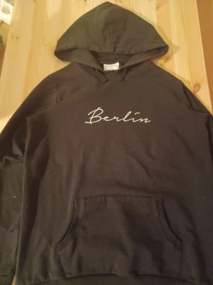 Svart hoodie med Berlin-tryck D-XEL - Svart hoodie från D-XEL med vit text 'Berlin' broderad på bröstet. Klassisk känguruficka framtill och justerbar huva. Tillverkad i mjukt bomullsmaterial som är skönt att ha på sig. Perfekt för en avslappnad streetwear-look.
