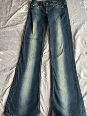 Blå utsvängda jeans med slitningar storlek 26/32 - 27/32 köptes för 400kr - Säljer ett par blå jeans med utsvängda ben och snygga slitningar framtill. Jeansen har klassisk femficksmodell, normal midja och dragkedja med knapp. Perfekta för dig som gillar retrostil och vill sticka ut med en cool look.