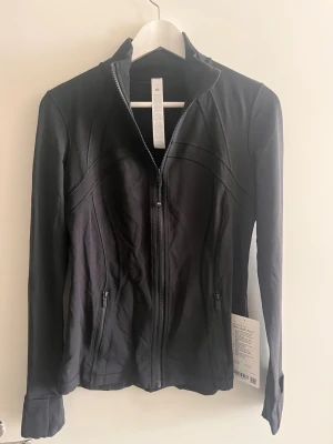 NY Lululemon Define Jacket Nalu  - Snygg svart träningsjacka från Lululemon med hel dragkedja, figurnära passform och två fickor med dragkedja. Tillverkad i ett stretchigt funktionsmaterial som andas. Perfekt för gymmet eller löprundan. Diskret logga på dragkedjan. 