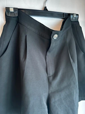 SHEIN svarta kostymshorts (Stl L) - Stilrena svarta kostymshorts från SHEIN i stl L (ca 40/42). Slät midja framtill med snygga pressveck, sidofickor samt melerad knapp. Gjorda i 100% polyester som håller färgen och är helt skrynkelfria. Toppenskick! Mått mätt plant: Midja: ca 39 cm (ej stretch) Längd: ca 41 cm Innerben: ca 11 cm Lårvidd: ca 35 cm Se sista bilderna för måttband & tvättråd!