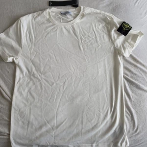 Vit t-shirt från Stone Island - Vit t-shirt från Stone Island med klassisk rund hals och korta ärmar. T-shirten har en enkel design med den ikoniska Stone Island-loggan på vänster ärm. Perfekt för en clean och stilren look.