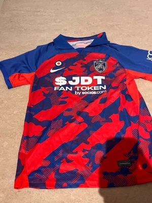 JDT matchtröja Nike röd/blå - Säljer en officiell JDT matchtröja från Nike i rött och blått med kamouflagemönster. Tröjan har klubbmärke och sponsortryck på bröstet samt klassisk krage. Perfekt för dig som vill stötta laget på match eller träning.
