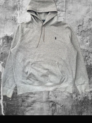Grå hoodie från Polo Ralph Lauren - Klassisk grå hoodie från Polo Ralph Lauren med broderad logga på bröstet. Hoodien har huva med dragsko, känguruficka och ribbade muddar vid ärmar och nederkant. Tillverkad i mjukt bomullsmaterial som är skönt att bära.