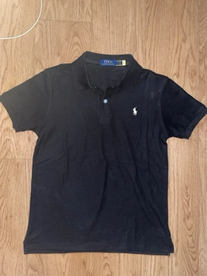 Svart pikétröja från Polo Ralph Lauren - Klassisk svart pikétröja från Polo Ralph Lauren med vit broderad logga på bröstet. Tröjan har korta ärmar, krage och knappar framtill. Tillverkad i mjuk bomull som känns skön mot huden. Perfekt för en clean och stilren look.