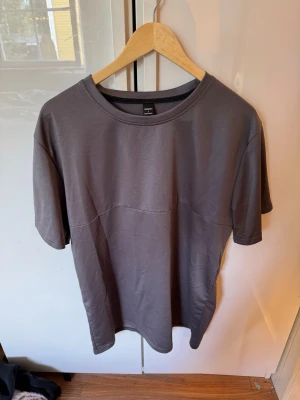 Mörkgrå basic t-shirt - En stilren mörkgrå t-shirt med rund hals och avslappnad passform. Tillverkad i mjuk bomull, perfekt för en enkel och clean look. T-shirten har korta ärmar och en diskret söm över bröstet som ger en modern touch.