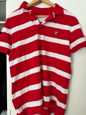 Vintage piké från American Eagle - Säljer en slim fit t-shirt från American Eagle med breda röda och vita ränder. Klassisk pikékrage och korta ärmar. Liten blå logga broderad på bröstet. Perfekt för dig som gillar en sportig och avslappnad stil.