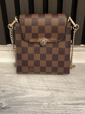 L och V axelväska i brunt rutmönster - Snygg axelväska från L och V i klassiskt brunt damier-rutmönster. Väskan har guldfärgad kedjerem och detaljer, samt ett elegant spänne framtill. Tillverkad i tåligt canvasmaterial med präglad logotyp på sidan. Kedjan kan man ta bort och ha väskan utan kedja. Den är perfekt för vardaglig användning där du kan få plats med många saker som kan behövas under dagen. Priset går att diskutera, flera bilder finns om det behövs. 