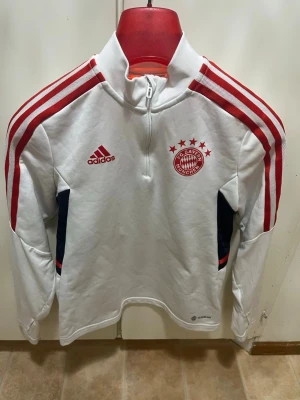 Bayern München träningströja - Snygg vit långärmad träningströja från Adidas med Bayern München-logga och röda detaljer. Tröjan har halv dragkedja, röda ränder på ärmarna och mörkblå inslag vid sidorna. Perfekt för fotbollsträning eller sportig stil.