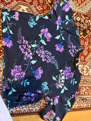 Blommig t-shirt med lila och blå toner - Snygg t-shirt med blommigt mönster i lila, blått och grönt på mörk botten. T-shirten har korta ärmar och rund halsringning. Perfekt för dig som gillar färg och mönster.