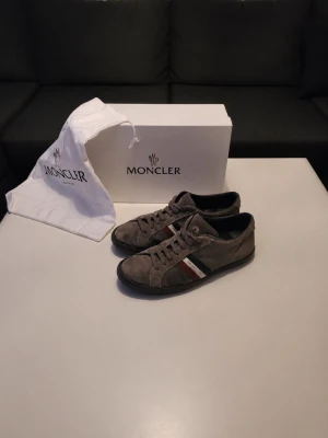 Moncler Skor - Snygga bruna sneakers från Moncler med klassiska ränder i rött, vitt och svart på sidan. Skorna har snörning och rund tå, samt en platt sula med Moncler-logga undertill. Utsidan är i mocka och insidan har vadderad skinnsula för extra komfort. Perfekt för dig som gillar stilrena detaljer.