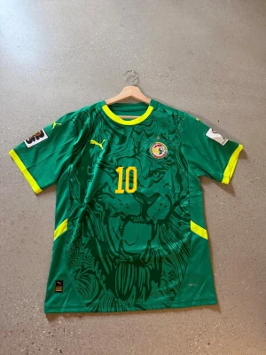 Senegal matchtröja Puma grön/gul - Grön Senegal matchtröja från Puma med gula detaljer och nummer 10 på bröstet och ryggen. Stort lejonmotiv på framsidan, Senegal-emblem på bröstet och texten 'Mané' på ryggen. Tillverkad i lätt funktionsmaterial, perfekt för fotboll. Helt ny replika 