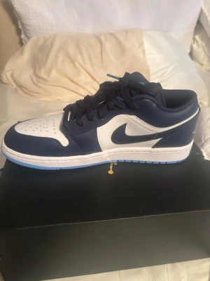 Nike Air Jordan 1 Low blå/vit - Snygga Nike Air Jordan 1 Low sneakers i marinblått och vitt läder. Klassisk siluett med perforerad tåbox, blå swoosh och matchande blå snörning. Yttersulan har en ljusblå detalj som ger extra stilpoäng. Perfekta för dig som gillar streetwear och ikoniska sneakers.