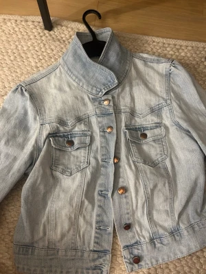 Ljusblå jeansjacka med hög krage - Trendig ljusblå jeansjacka med krage och snygga bronsfärgade knappar. Jackan har två bröstfickor med lock och knapp, samt markerade sömmar för en cool look. Perfekt att slänga över en hoodie eller t-shirt för en avslappnad stil.💙💙💙Den är liten i modellen , sitter ungefär som S men står storlek 40. 💙