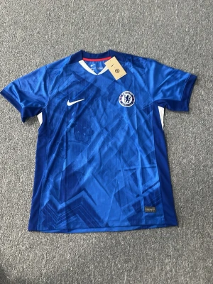 Chelsea FC blå matchtröja Nike - Chelsea FC matchtröja i blått med vita och röda detaljer. Tröjan har klubbmärke på bröstet, Nike-logga och ett grafiskt mönster över hela tyget. Tillverkad i ett lätt och ventilerande material, perfekt för fotboll eller träning.