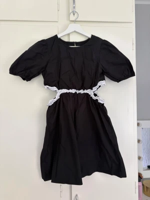 Svart kortklänning med cut-outs - Svart kortklänning med puffärmar och vita spetsdetaljer runt midjan som skapar snygga cut-outs. Klänningen har rund halsringning och dragkedja i ryggen. Perfekt för dig som vill ha en unik och trendig look. Små blå fläckar på den vita delen