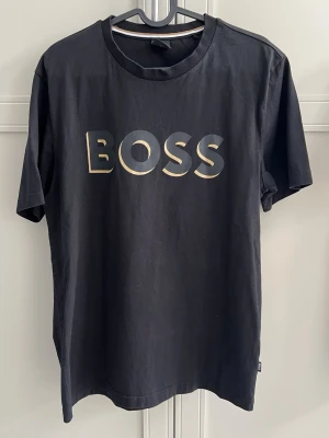 Svart BOSS t-shirt med tryck - Snygg svart t-shirt från BOSS med stort BOSS-tryck i på bröstet. Klassisk rund halsringning med en diskret rand i vitt och guld på insidan av kragen. Perfekt för en stilren och modern look.