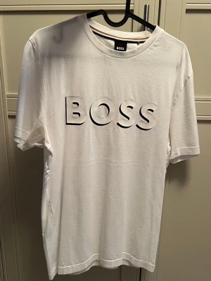 Vit BOSS t-shirt med tryck - Vit t-shirt från BOSS med tryck på bröstet. Klassisk rund halsringning och korta ärmar. Tillverkad i mjuk bomull för en skön och avslappnad känsla. Perfekt för en clean och stilren look.