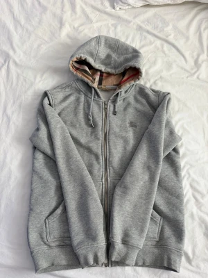 Grå zip hoodie från Burberry - Säljer en grå zip hoodie från Burberry med klassiskt rutigt foder i huvan och dragsko. Tröjan har två fickor framtill och diskret Burberry-logga på bröstet. Perfekt för dig som gillar stilrena och bekväma plagg med en touch av lyx.