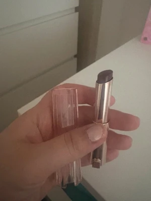 Lancome läppstift - Snyggt mörklila läppstift i en elegant, genomskinlig rosa hylsa med gulddetaljer. Läppstiftet har en klassisk rundad form och ger en djärv färg till läpparna. Perfekt för dig som vill sticka ut med en unik look.