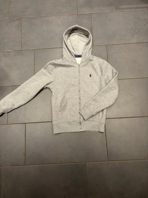 Grå zip hoodie från Polo Ralph Lauren - Snygg gråmelerad hoodie med dragkedja från Polo Ralph Lauren. Klassisk design med huva, två fickor fram och den ikoniska lilla blå loggan broderad på bröstet. Tillverkad i mjukt bomullsmaterial som är skönt att ha på sig.