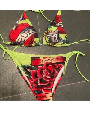 Färgglad bikini med rosor och text - Säljer en färgstark trekantsbikini med knyt i sidorna och över nacken. Bikinin har ett coolt tatueringsinspirerat mönster med röda rosor och text på både överdel och underdel. Basen är limegrön med inslag av svart, rött och blått. Perfekt för dig som vill sticka ut på stranden!