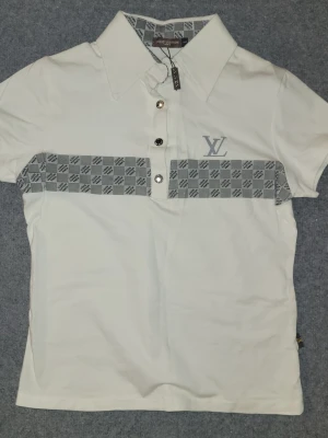 T-shirt med mönster - Vit t-shirt med grått mönster över bröstet och knappslå med tre knappar. Klassisk krage och korta ärmar. Snygg och stilren design som sticker ut. Ettikett kvar.