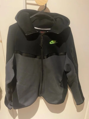 Nike tech fleece tracksuit svart och grå med grön logo - Snygg hoodie från Nike i svart och mörkgrått med dragkedja framtill och huva. Har en grön Nike-logga på bröstet och en ficka med dragkedja på ärmen. Tillverkad i mjukt material, perfekt för en sportig och avslappnad stil.