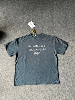 Grå Acne Studios t-shirt - Snygg grå t-shirt från Acne Studios med tryck 'STOCKHOLM 1996' på bröstet. Klassisk passform med korta ärmar och rund halsringning. Perfekt för en avslappnad och trendig look.