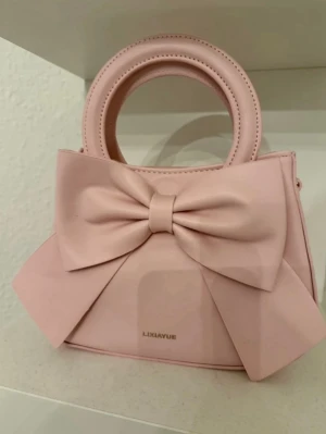 Ljusrosa handväska med stor rosett - Supersöt ljusrosa handväska med en stor rosett på framsidan. Väskan har rundade handtag och avtagbar axelrem i matchande färg. Perfekt för dig som gillar gulliga accessoarer och passar verkligen alla åldrar! 💕