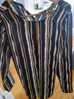 Randig blus med knytband bak - Snygg blus med svarta, beige och vita vertikala ränder. Blusen har lång ärm och ett dekorativt knytband i ryggen. Tillverkad i ett tunt, lätt och något transparent material som ger en luftig känsla. Perfekt för dig som gillar trendiga mönster och detaljer.