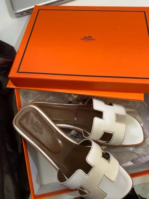 Vita slip-in sandaler med H-design - Säljer ett par trendiga vita sandaler med öppen tå och breda remmar i H-form. Sandalerna har platt sula och är tillverkade i slätt konstläder. Perfekta för sommaren och ger en clean look till din outfit.