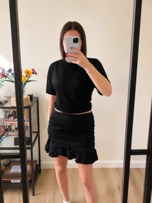 Svart kortkjol från Vero Moda - Säljer en svart kortkjol från Vero Moda med volangkant och rynkade detaljer. Kjolen har en figurnära passform och är tillverkad i ett mjukt material som känns bekvämt. Perfekt att styla med en enkel t-shirt eller crop top.