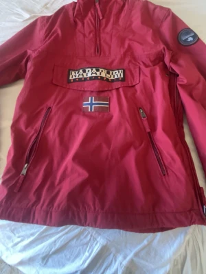 Röd vindjacka från Napapijri - Röd vindjacka från Napapijri med dragkedjor på sidorna, stor ficka framtill med logga och norsk flagga. Jackan har en patch på ärmen och är tillverkad i ett lätt, vindtätt material. Perfekt för blåsiga dagar och streetwear-stil.