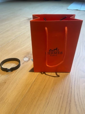 Svart armband från H - Snyggt svart armband från H i metall med klassisk H-detalj framtill. Armbandet har en stilren och modern design som passar perfekt till din outfit. Levereras med original orange H-påse.