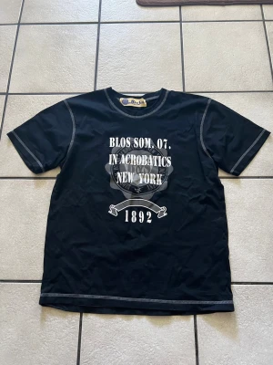 Svart t-shirt med tryck från Blos Som storlek M/L - Cool svart t-shirt med vit text och tryck på bröstet där det står 'BLOS SOM. 07. IN ACROBATICS NEW YORK 1892'. T-shirten har kontrastsömmar och klassisk rund hals. Perfekt för en avslappnad streetwear-look.