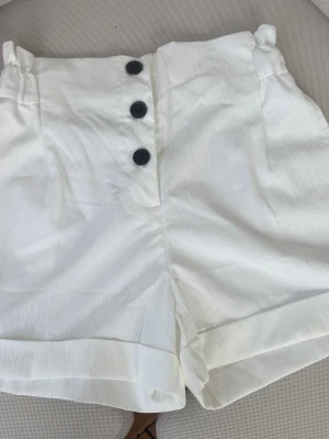 Vita högmidjade shorts med knappar - Stilrena vita shorts med hög midja och tre svarta knappar framtill. Shortsen har resår i midjan baktill och uppvikta benslut för en modern look. Tillverkade i ett lätt tyg som passar perfekt till varma dagar.